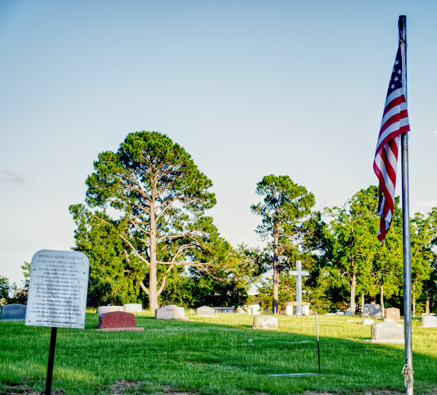A historical marker and American flag stand reamain between Cedar Memorial Gardens and the Black cemetery in Mineola, with a cross in the background on the other side. The fence is no longer present.